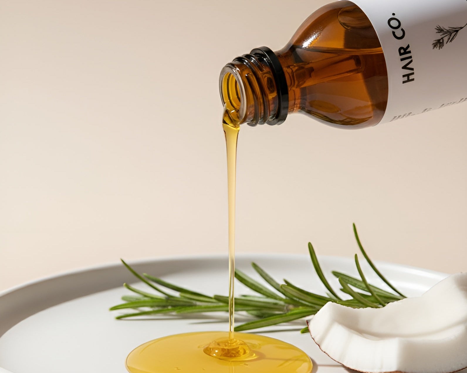 Hair oil being poured from a bottle labeled 'Hair Co.' onto a white plate with rosemary and coconut.